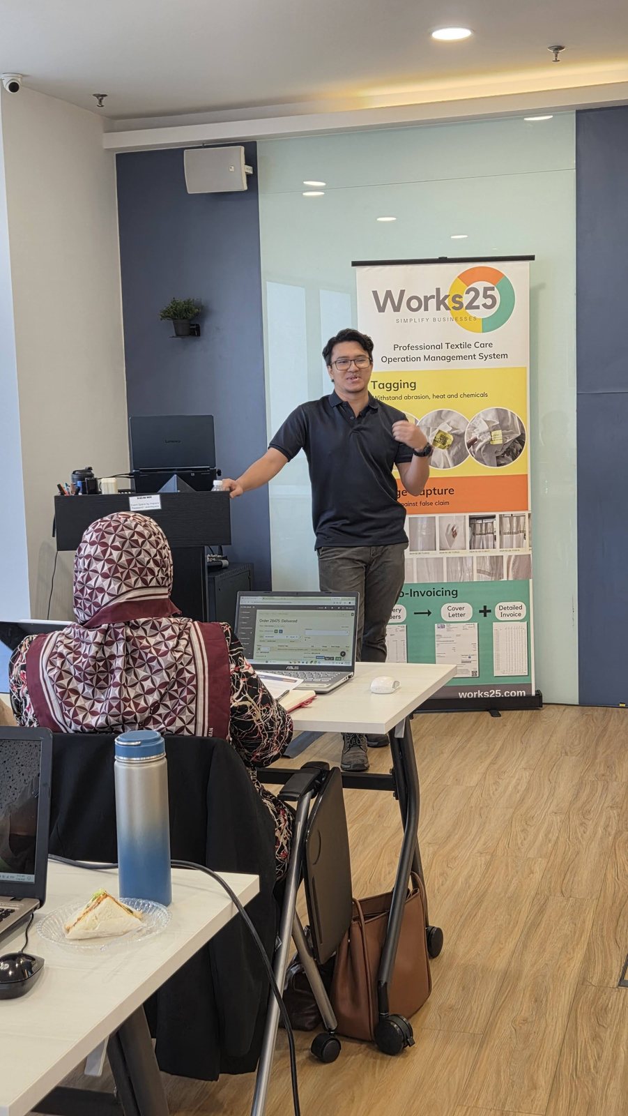 Pembentang berdiri di hadapan peserta di sebelah sepanduk Works25